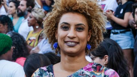 Marielle Franco smiling. Her hair is short, blond and curly. She is wearing a floral blouse and a pair of bright blue earings. 