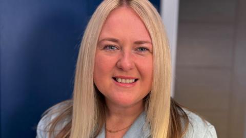 Head-and-shoulders portrait of Lesley with long light-coloured hair, wearing a light blue collared top and a thin necklace, looking toward the camera against a softly blurred indoor background with dark blue and neutral tones.