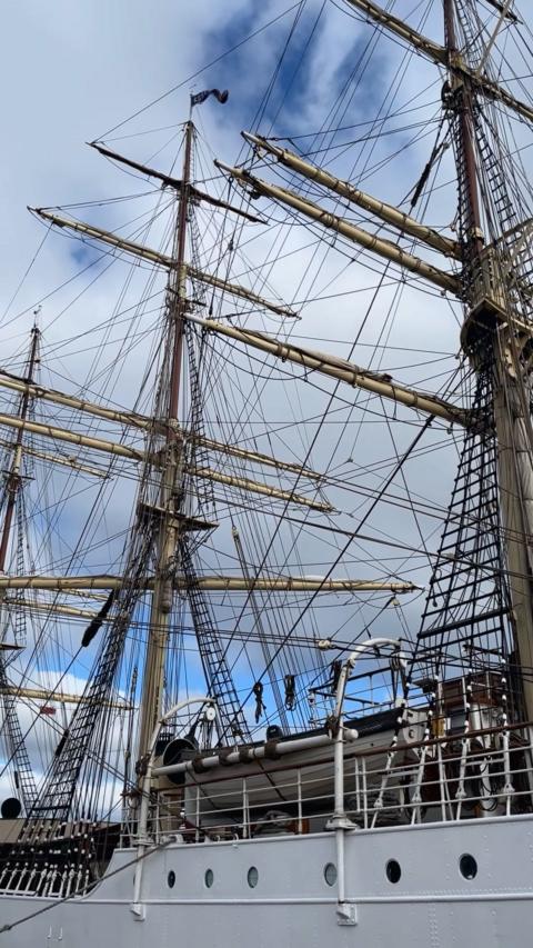 A white tall ship has three masts, its sails are tied up, against a cloudy sky