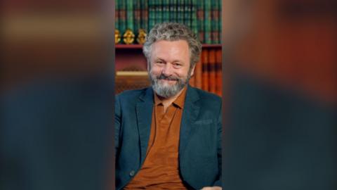 A man sits in a chair with lots of books on a shelf behind him. He is smiling and is wearing a blue blazer and mustard coloured polo top. 