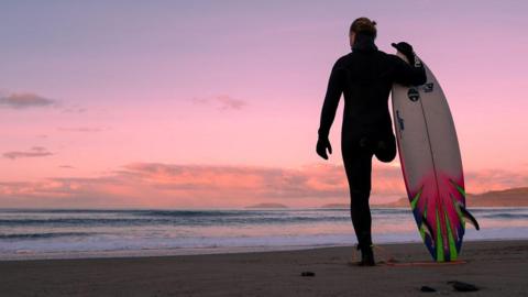 Surfer in a full body black wetsuit and gloves. He has one leg and he stands on a beach at colourful cool pink sunset. He has his back to the camera with the sea in front and the vast sky beyond. He's holding a white and shocking pink and green surfboard with its tip pointing skyward and the other end resting on a sandy beach.
