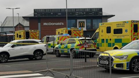 Three ambulances can be seen outside a McDonald's branch. Two response vehicles can also be seen, with a police car in the foreground. Two metal fences cordon the area off, with a white car also seen to the side. 