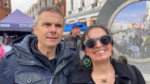 Neil and Ana Ball are looking at the camera from Ipswich town centre, in front of the "portal" to Brazil. She has dark hair which is tied back and sunglasses on. He has short, grey hair and is wearing a blue top and jacket.