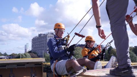 A man in blue top and white shorts and orange hat prepares to abseil down a football stadium stand. Next to him is a woman in an old gold football shirt and orange helmet