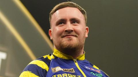Luke Littler, pictured in March 2026, wearing a blue and yellow jacket during a darts tournament