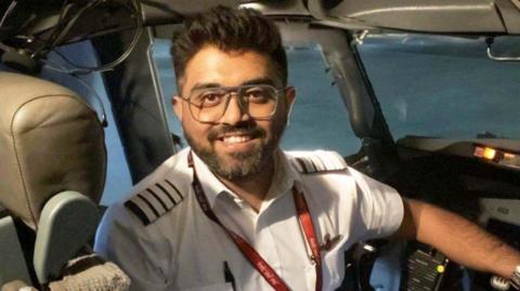 Indian pilot Smit Machchhar seen smiling into a camera as he sits in the cockpit of a plane. He is wearing glasses, a white shirt and has a pen tucked in his shirt pocket.