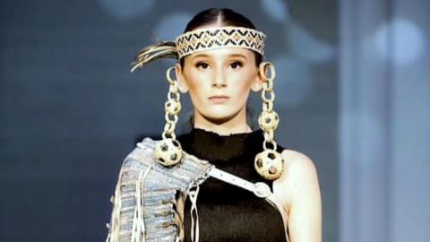 Gracie is modelling on the catwalk at London Fashion week wearing a headband with very large chunky loops coming from the top of her ears and falling below her shoulders.  Gracie has a black round neck sleeveless dress with a garment thrown over one shoulder. 