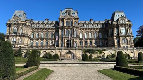 A large building in the French Renaissance style of architecture - four storeys with large windows, projecting bays, and mansard roofing. Stairs on either side lead up the the central entrance. In front is a formal garden with a wide gravel path, patches of lawn, and clips shrubbery.