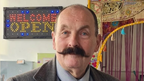 A balding man with a dark handlebar moustache is wearing a tweed jacket and standing in front of a sign with the words welcome open. 