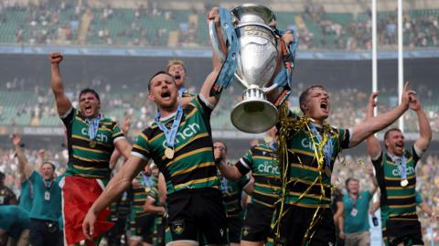 Fraser Dingwall (left) holds the Prem trophy aloft in his left arm and Fin Smith (right) holds it in his left, with his right arm held up to his side and finger pointing. The pair are walking on the pitch at the Allianz Stadium with their mouths open after winning the Prem final