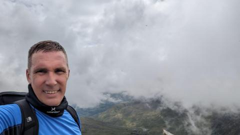A man in a blue top is standing on Mount Snowdon, the view is visible on the right of the photo