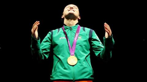 Katie Taylor looks up to the sky with her hands up and gold medal around her neck