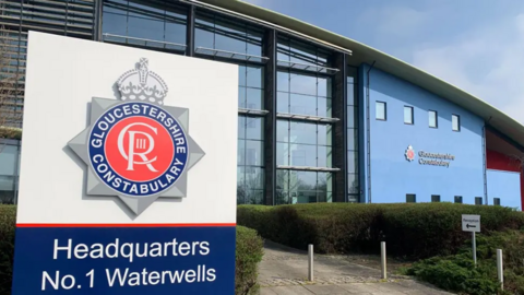 The exterior of Gloucestershire Police headquarters. It is a blue building with a curved roof, and the force's logo is on a white and blue sign in the foreground