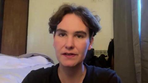 Robyn Lewis is sat down in a hotel room. She is sat next to an unmade bed and a set of grey curtains. She has short brown hair and is wearing a black t-shirt. A door can be seen behind here along with a light yellow wall.