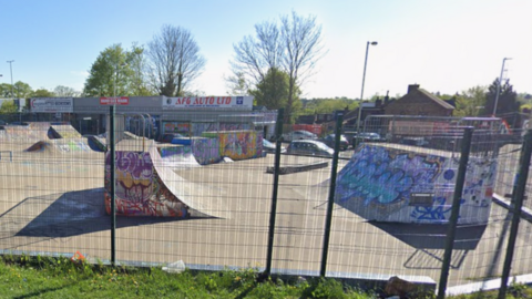 A skatepark, with fencing round it, showing graffiti on a large number of ramps. There is metal railing at the front of the photo and trees in the distance. 