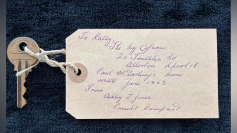 A silver house key with a paper label attached to it with string through the hole of the key. The paper label has old-fashioned handwriting on it which reads "To Kathy, the key from 20 Forthlin Road, Allerton, Liverpool 18. Paul McCartney's home until June 1963, from Ashley J Jones, present occupant". The key and label have been placed on navy blue background material.