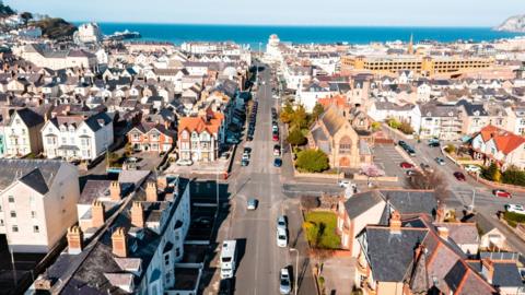 Drone photo of Llandudno town, houses and streets are visible, with cars driving on the roads. In the distance the sea is visible. 