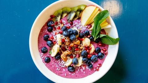 A stock photo of a white bowl with purple acai paste and fruits including berries, banana, pomegranate seeds, kiwi, and mint leaves and nuts 