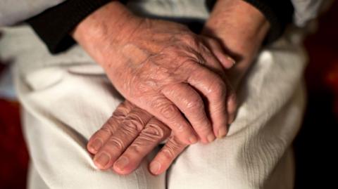 An older person poses with their hands folded on their lap. 