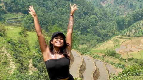 Juliana Marins raising both hands in the air, in a black cap and black tube top. In the background are green paddy fields.