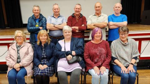 Five women are sat on school chairs and five men stood behind with their arms folded.