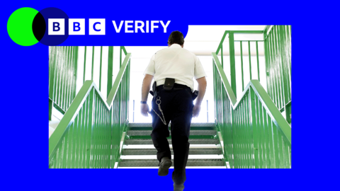 A prison officer wearing a white shirt and black trousers walks up a flight of stairs with his back to the camera.
