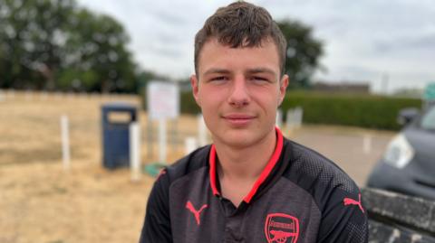 A teenage boy in a black Arsenal shirt, with red trim and badge, sits on a bench at the edge of a football pitch.