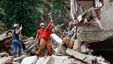 Two men and a woman stand on top of rubble next to a collapsed building. They are wearing hard hats and one of them is wielding a sledgehammer as they search for survivors.