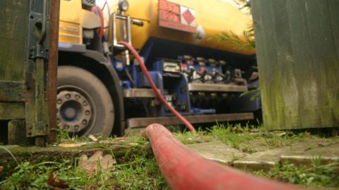 A large red and blue truck parked up delivering domestic heating oil through a red pipe, seen at the front of the shot. 
