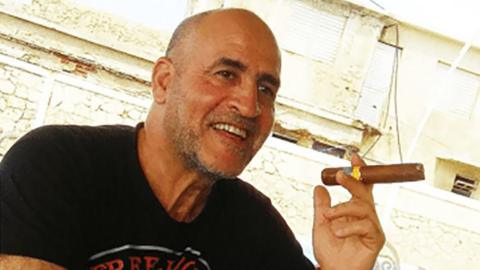 Daniel Siad, a man with a shaved head and stubble, wearing a black T-shirt, sits outside in what appears to be a sunny street, smiling with a large cigar raised in his hand.