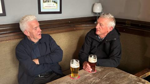 Two men with silver/white hair and wearing dark clothing sitting at a pub table with two pints of beer in front of them