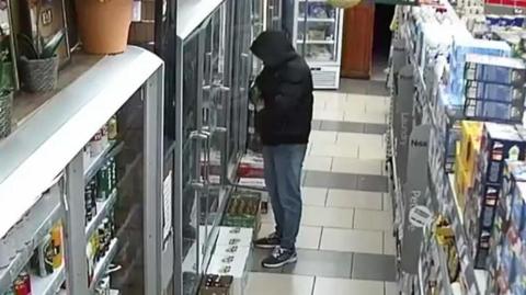 CCTV of a man standing in a shop. He is wearing a black jacket with a hood that covers his face and blue jeans. He appears to be tucking something into the front of the jacket