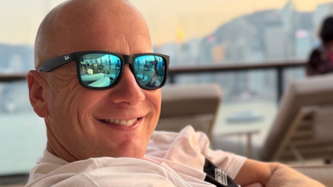 A photo of a man wearing black glasses with blue reflective lenses, grinning at the camera.