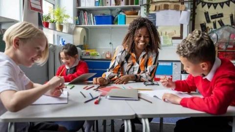 A group of kids around a set of table that are pulled together with a teacher, using pens and paper. 