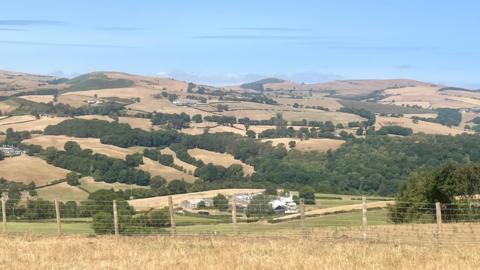 The view from Nantglyn, Denbigshire of an agricultural landscape covered with parched fields. There is a fence in the foreground, with dry hills in the background. 
