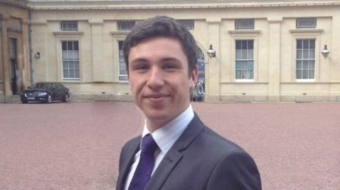 George Elliott picture at Buckingham Palace in London. He is wearing a black suit, white shirt and purple tie, looking at the camera and smiling.