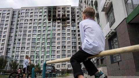 A little boy in a white embroidered shirt and black trousers looks at a building damaged in a Russian air attack, in Kyiv