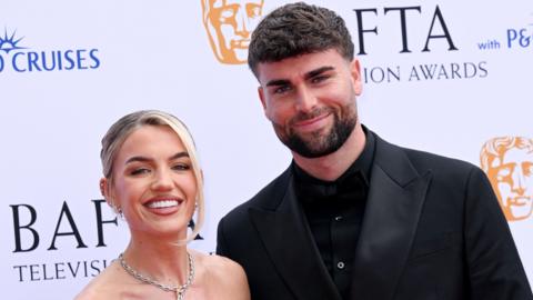 Molly Smith and Tom Clare pose together. Molly is wearing a silver necklace and off-the-shoulder dress and Tom is wearing a black suit over a black shirt.  