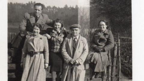 A black and white image of two men and three women in long coats.
