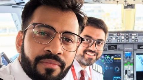 Indian pilot Smit Machchhar and his friend  Captain Dharav Gosalia smiling into a camera as they sit in the cockpit of a plane. 
