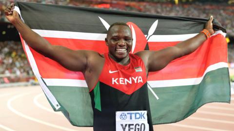 Julius Yego, visible from waist up, smiles broadly as spreads his arms wide to raises a Kenyan flag wide behind himself after winning gold in the javelin at the 2015 World Athletics Championships in Beijing. He wears a red and black vest with the word 'Kenya' written in white text across the chest and a white bib with his name is pinned below. Yego stands on a terracotta running track while a packed stand is blurred in the background