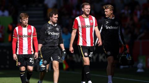 Nathan Collins, in Brentford's red-and-white striped kit, black shorts and black socks, grimaces as he walks off the pitch flanked by two physios