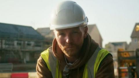 A builder wearing hi-vis clothing and a white hard hat looks into the camera