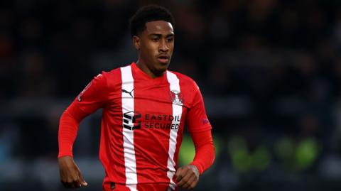 Zech Obiero running, head and torso shot, looking forward, wearing red and white home shirt of Leyton Orient.