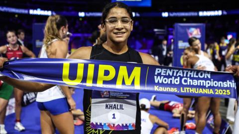 Farida Khalil, wearing clear glasses and a black vest with a bib on it bearing her surname and the number one, holds a banner reading Pentathlon World Championships, Kaunas, Lithuania 2025 across her chest. Several other athletes can be seen out of focus behind her in an indoor arena, either standing up or lying on the floor