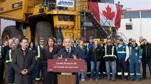 Mark Carney stands before a podium with a sign that reads in French and English: 'Canada Strong for All'. 
He is flanked by Alberta Premier Danielle Smith, and behind him are oil workers in hard hats and work vests. A large piece of yellow equipment is hung with a Canadian flag. 