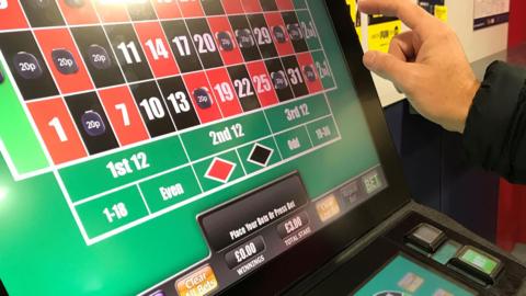 A man's hand is pictured about to press the brightly lit screen of a slot machine. The screen shows a series of red and black squares, some of which are filled with the price of bets placed. Details of stakes and winnings are also pictured. 