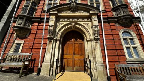 The outside of the entrance to the House of Keys, which is a red brick building with large wooden double doors with an ornate surrounding.