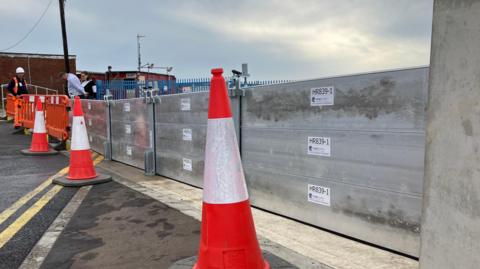 Three cones positioned in front of a large metal barrier spanning from the left to the right of the image. 