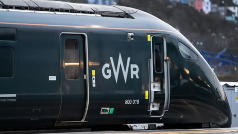 A dark green Great Western Railway train with a sloped nose passes along a platform.